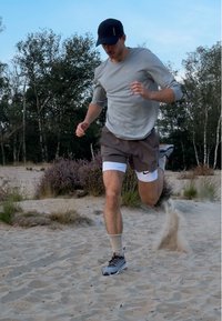 Maglietta grigia a maniche lunghe, pantaloni corti marroni, mutande bianche e sneakers grigie. In corsa sulla sabbia con erba e cespugli sullo sfondo.