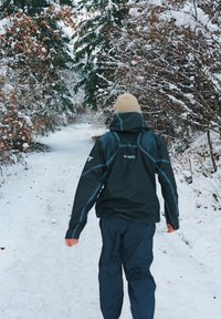 Must veekindel jope siniste aktsentidega, kantud tumedate pükstega, kõndides lumega kaetud rajal, mida ääristavad lumised puud ja taimestik.