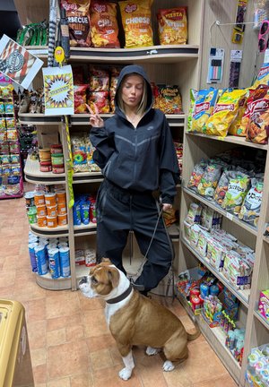 Chaqueta negra con cremallera y capucha, pantalones a juego, y un perro marrón claro con correa. Estantes de la tienda llenos de snacks y bebidas de fondo.