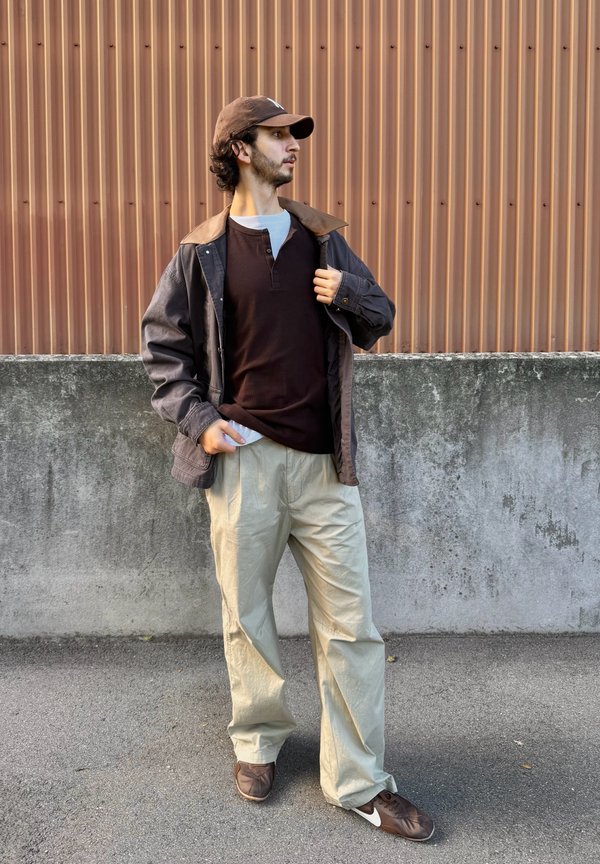 Un hombre con una gorra y una chaqueta marrones está junto a una pared de hormigón, mira hacia un lado y sujeta la solapa de su chaqueta.