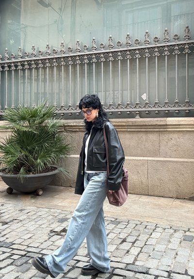 Chaqueta de cuero negra, camisa blanca, jeans de mezclilla azul claros, zapatos negros gruesos y un bolso rosa. En un entorno urbano con una planta de palma.