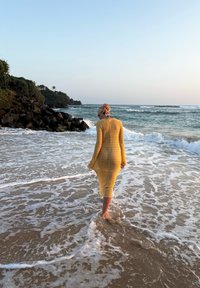 Personne en robe jaune debout pieds nus dans l'eau peu profonde de l'océan près d'un rivage rocheux au coucher du soleil.