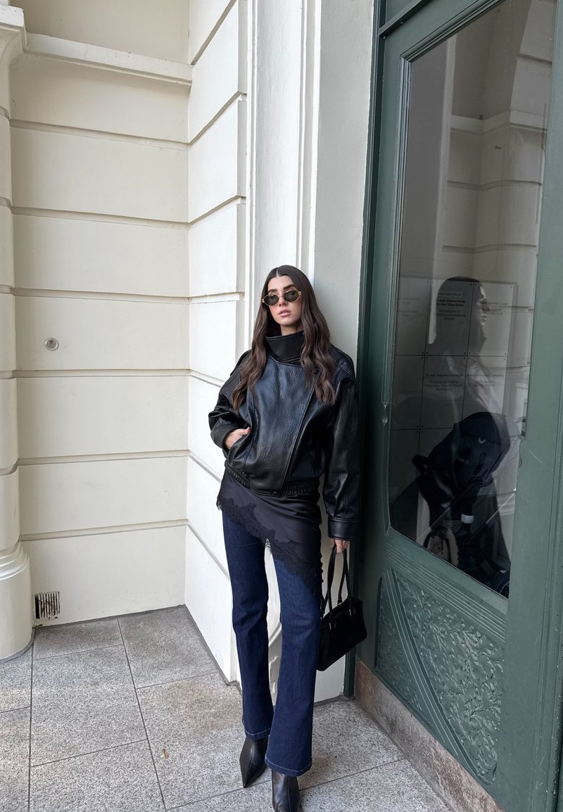 Femme aux longs cheveux noirs portant des lunettes de soleil, une veste en cuir noire, un jean foncé, des bottes noires, tenant un sac à main noir, appuyée contre un bâtiment.