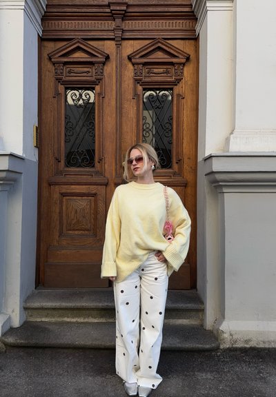 Mujer con suéter amarillo oversize, pantalones blancos con lunares negros, gafas de sol y zapatos plateados, de pie frente a unas puertas dobles de madera.