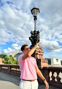 Pink polo shirt, white shorts, and a bronze lamppost featuring ornate figures. Sky with clouds, subtle textures in clothing and background.