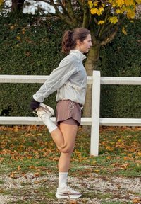 Atletyczna kobieta rozciąga się, trzymając stopę z tyłu, ubrana w jasnoszarą kurtkę, brązowe szorty oraz białe skarpetki i buty do biegania.