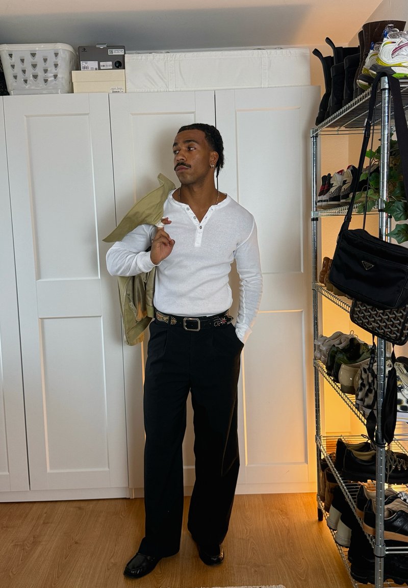 Man in wit shirt met lange mouwen en zwarte broek houdt een groene jas over zijn schouder, staat bij witte kasten en een schoenenrek.