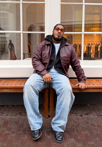 Chaqueta de cuero marrón con forro de forro polar negro, usada sobre una camisa blanca. Vaqueros de pierna ancha azul claro y zapatos negros. Banco de madera y ventana de fondo.