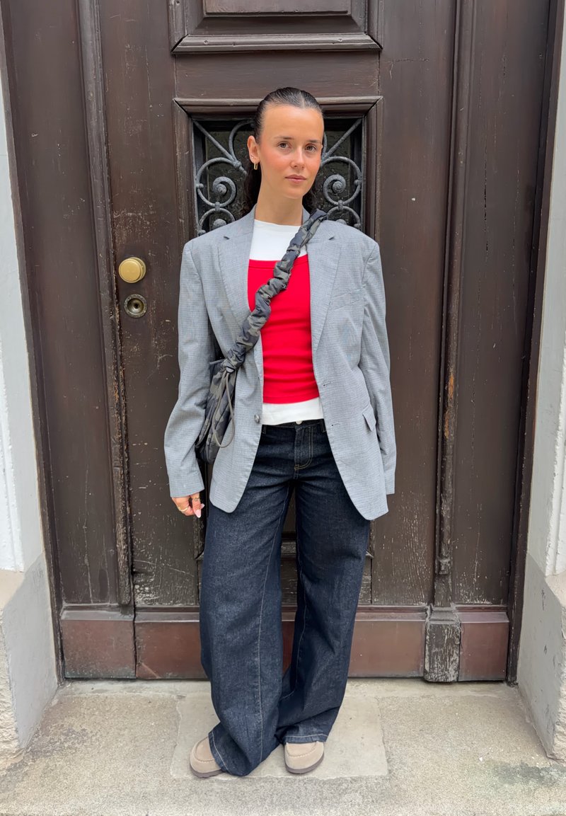 Femme debout devant une porte en bois sombre, portant un blazer gris, des hauts superposés rouges et blancs, un jean large noir, des chaussures beige et un sac bandoulière.