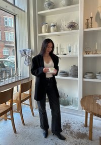 Blazer noir sur un haut en dentelle blanche, associé à un jean noir à jambes larges et des bottines. Tenant une tasse à thé, entouré de verrerie et de céramiques.