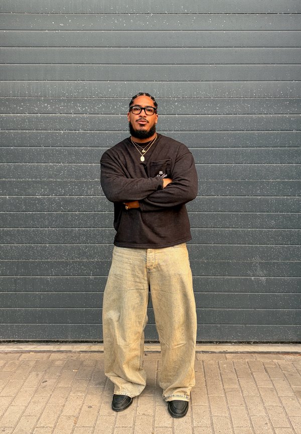 Homme aux cheveux tressés et lunettes, debout les bras croisés, portant un pull noir, un pantalon large beige et des chaussures noires devant un mur gris.