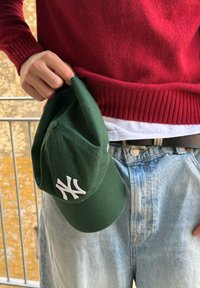 Roheline pesapallimüts valge tikkitud logoga, hoitakse ühes käes, kantud punase kampsuni ja helesiniste teksadega. Omaduseks on ümar ääreline.