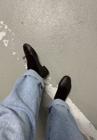 Personne portant un jean bleu clair retroussé, des chaussettes foncées et des ballerines noires, debout sur un sol gris avec des marquages blancs.