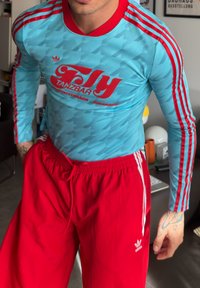 Άνδρας που φοράει ανοιχτό μπλε μακρυμάνικο πουκάμισο με κόκκινη ρίγα και το λογότυπο "Tanzbar Fly" και φωτεινά κόκκινο παντελόνι Adidas με λευκές ρίγες.