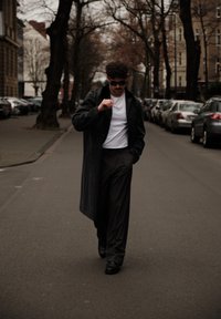 Un homme portant des lunettes de soleil et un long manteau noir marche dans une rue de la ville bordée de voitures garées et d'arbres sans feuilles.