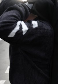 Personne portant un pull noir duveteux avec des motifs géométriques blancs, bras plié et main gantée de noir près de la tête.