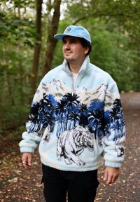 Fleece-pullari vaaleansinisenä, palmupuukuvioilla ja tiikerikuviolla. Vetoketjulla varustettu kaulus, teksturoitu kangas, pitkät hihat, mustien housujen kanssa.
