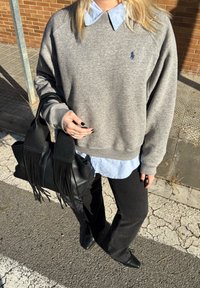 Personne portant un sweat-shirt gris par-dessus une chemise bleue à col, pantalon noir, chaussures noires, tenant un sac à main en cuir noir à franges sur le trottoir.