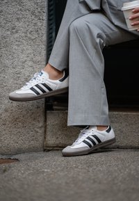 Vita lädersneakers med svarta ränder, grå gummisula och rund tå. In tillsammans med grå byxor, placerade på en stenyta.