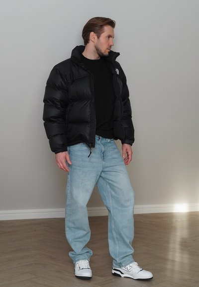 Chaqueta de plumas negra con cuello alto y cremallera frontal, combinada con jeans de mezclilla azul claro y zapatillas blancas con detalles en negro.