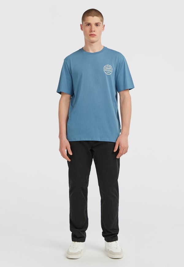 Camiseta de algodón azul con mangas cortas y cuello redondo. Presenta un estampado gráfico en el pecho. Combinada con pantalones negros y zapatillas blancas.