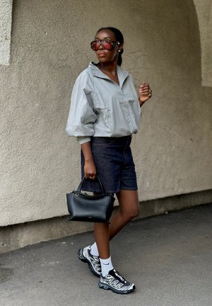 Mujer con chaqueta clara de gran tamaño, pantalones cortos oscuros, zapatillas deportivas, calcetines blancos, grandes gafas de sol rojas, llevando bolso negro, de pie frente a una pared texturizada.