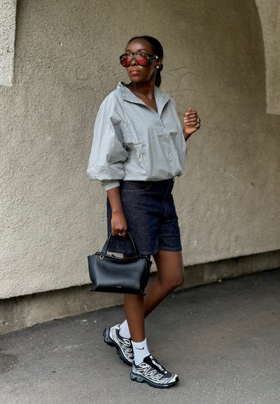 Mujer con chaqueta clara de gran tamaño, pantalones cortos oscuros, zapatillas deportivas, calcetines blancos, grandes gafas de sol rojas, llevando bolso negro, de pie frente a una pared texturizada.
