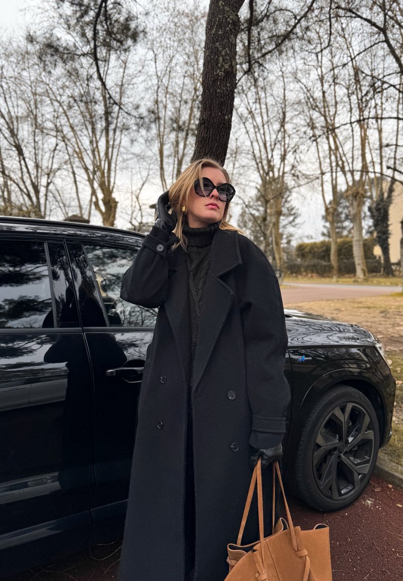 Manteau en laine noir surdimensionné, pull en maille noir, gants noirs. Sac cabas en cuir marron. Porte de grandes lunettes de soleil noires, debout à côté d'une voiture noire.