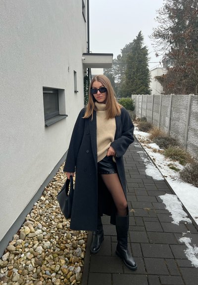 Mujer con abrigo negro, suéter de cuello alto beige, pantalones cortos de cuero negro, botas negras y gafas de sol, de pie en un camino pavimentado junto a un edificio blanco.