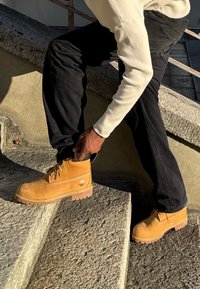 Botas de tobillo amarillas de ante con puntera reforzada, diseño de cordones y suela resistente. Llevadas por una persona en escalones de piedra.