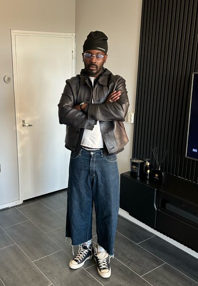 Chaqueta de cuero marrón sobre una camisa blanca, culottes anchos de denim y zapatillas negras. Ambientado en un interior moderno con detalles en tonos oscuros.