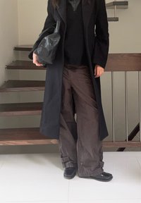 Fekete hosszú kabát, sötétbarna, széles szárú, mandzsettás nadrág, fekete bőr táska és fekete cipő, háttérben fa lépcsővel.