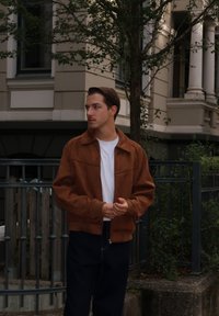 Giacca bomber in suede marrone con colletto, zip frontale e due tasche, indossata sopra una t-shirt bianca e jeans scuri. Dettagli in un contesto urbano.