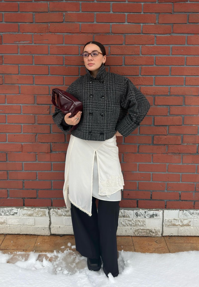 Femme portant une veste à carreaux sombre, jupe asymétrique blanche sur un pantalon noir, tenant une pochette bordeaux brillante, debout près d'un mur en briques rouges sous la neige.