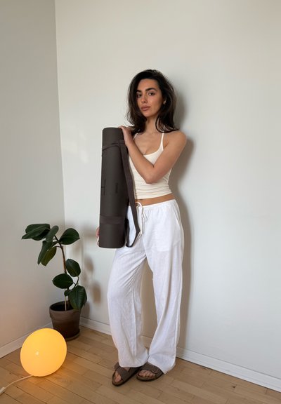 Mujer con top y pantalones blancos sosteniendo una esterilla de yoga enrollada, de pie junto a una planta en maceta y una lámpara de pie redonda y luminosa contra una pared lisa.