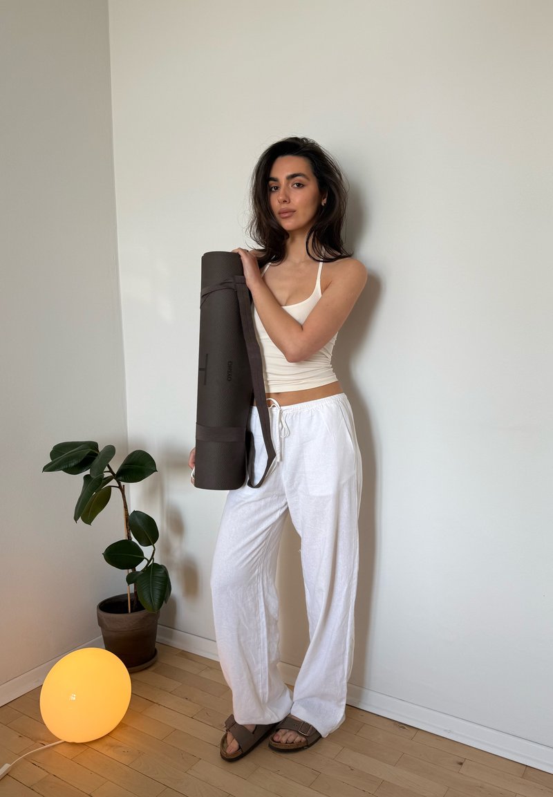 Femme en haut blanc et pantalon tenant un tapis de yoga roulé, debout près d'une plante en pot et d'une lampe de sol ronde lumineuse contre un mur uni.