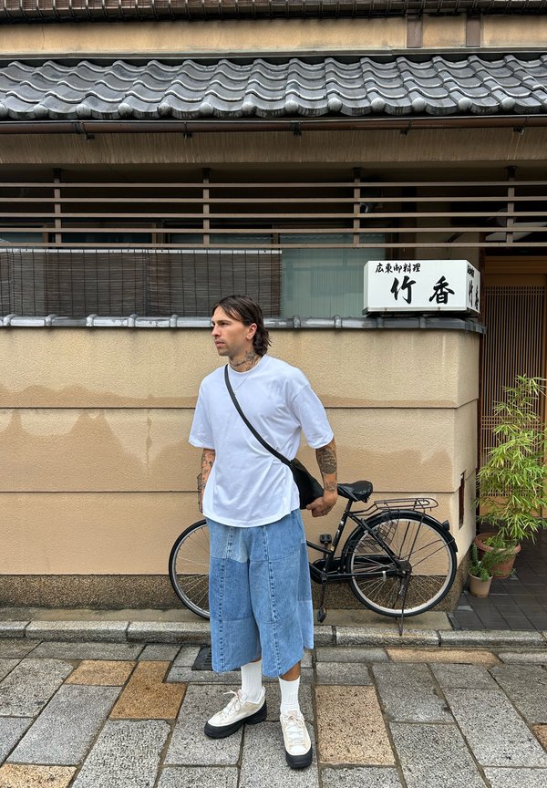 Hombre con tatuajes que viste una camiseta blanca y pantalones cortos de mezclilla de retales, de pie sobre un pavimento de piedra sosteniendo una bicicleta negra cerca de una pared beige con un cartel japonés.