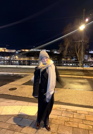 Persona con abrigo de invierno, bufanda y sombrero de pie en una calle pavimentada durante la noche, con luces de la ciudad y un río de fondo.