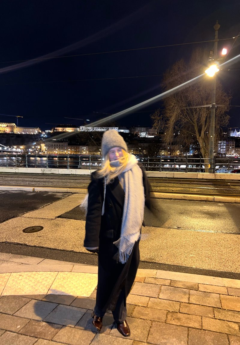 Personne en manteau d'hiver, écharpe et chapeau debout sur une rue pavée la nuit, avec les lumières de la ville et une rivière en arrière-plan.