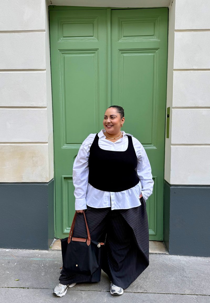 Femme en gilet noir, chemise blanche et pantalon large rayé sourit avec un sac à main noir devant des portes doubles vertes dans une rue de la ville.
