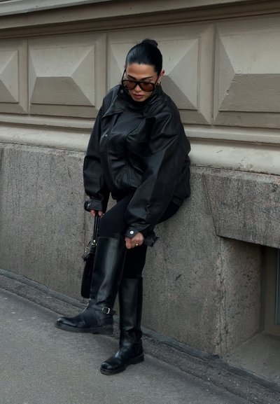 Mujer con chaqueta de cuero negra y botas ajustándose la bota mientras está de pie en una acera de la ciudad junto a un muro de piedra.
