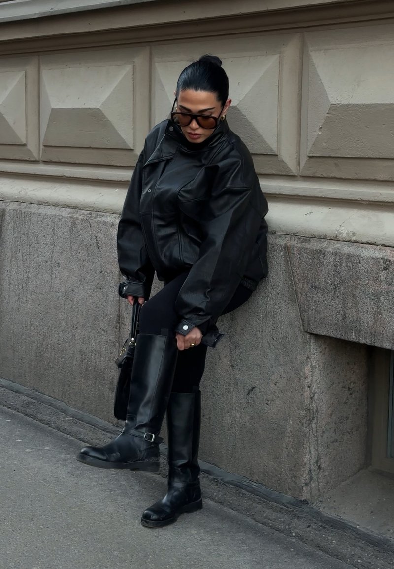 Femme en veste en cuir noire et bottes ajustant sa botte tout en se tenant sur un trottoir en ville à côté d'un mur en pierre.