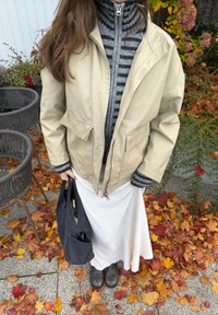 Beige zipjacka över en grå randig polotröja, tillsammans med en vit strukturerad kjol och svarta skor, hållande en mörk väska bland höstlöven.