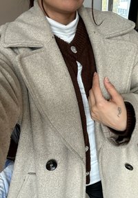 Person som bär en beige ullkappa över en brun knappad tröja och en vit polotröja, med en liten tatuering på vänster handled.