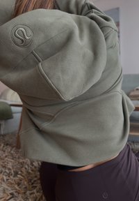 Hanorac verde măsliniu cu textură canelată, având un logo rotund pe mânecă și o croială scurtă, asortat cu leggings negri cu talie înaltă.