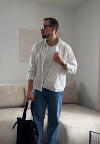 Άντρας που φοράει γυαλιά ηλίου, ριγέ πουκάμισο, λευκή μπλούζα και τζιν, κρατώντας μια μαύρη τσάντα, στέκεται δίπλα σε μια μπεζ καναπέ σε ένα μινιμαλιστικό σαλόνι.