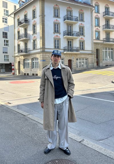 Gabardina larga beige sobre suéter azul marino con texto en blanco, camisa blanca con cuello y pantalones plisados grises claros. Zapatos negros. Entorno urbano.