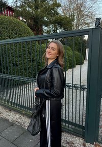 Blouson en cuir noir avec des détails de fermeture éclair argentée, associé à une tenue noire avec trois rayures blanches. Sac à main texturé et fond de haie verte.
