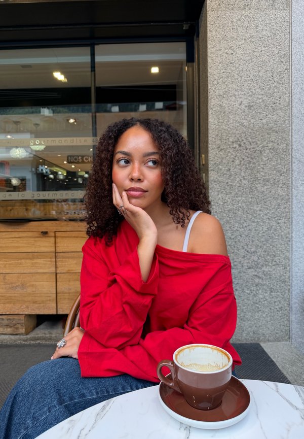 Une femme vêtue d’un haut rouge est assise à l’extérieur d’un café, le menton posé sur une main, à côté d’une tasse de café.