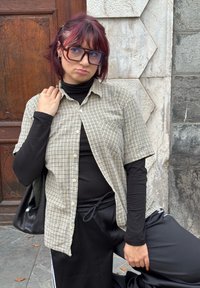 Košile s krátkým rukávem s károvaným vzorem v béžové a hnědé barvě, nošená přes černý rolák. Má na sobě velké hranaté brýle a černou tašku.